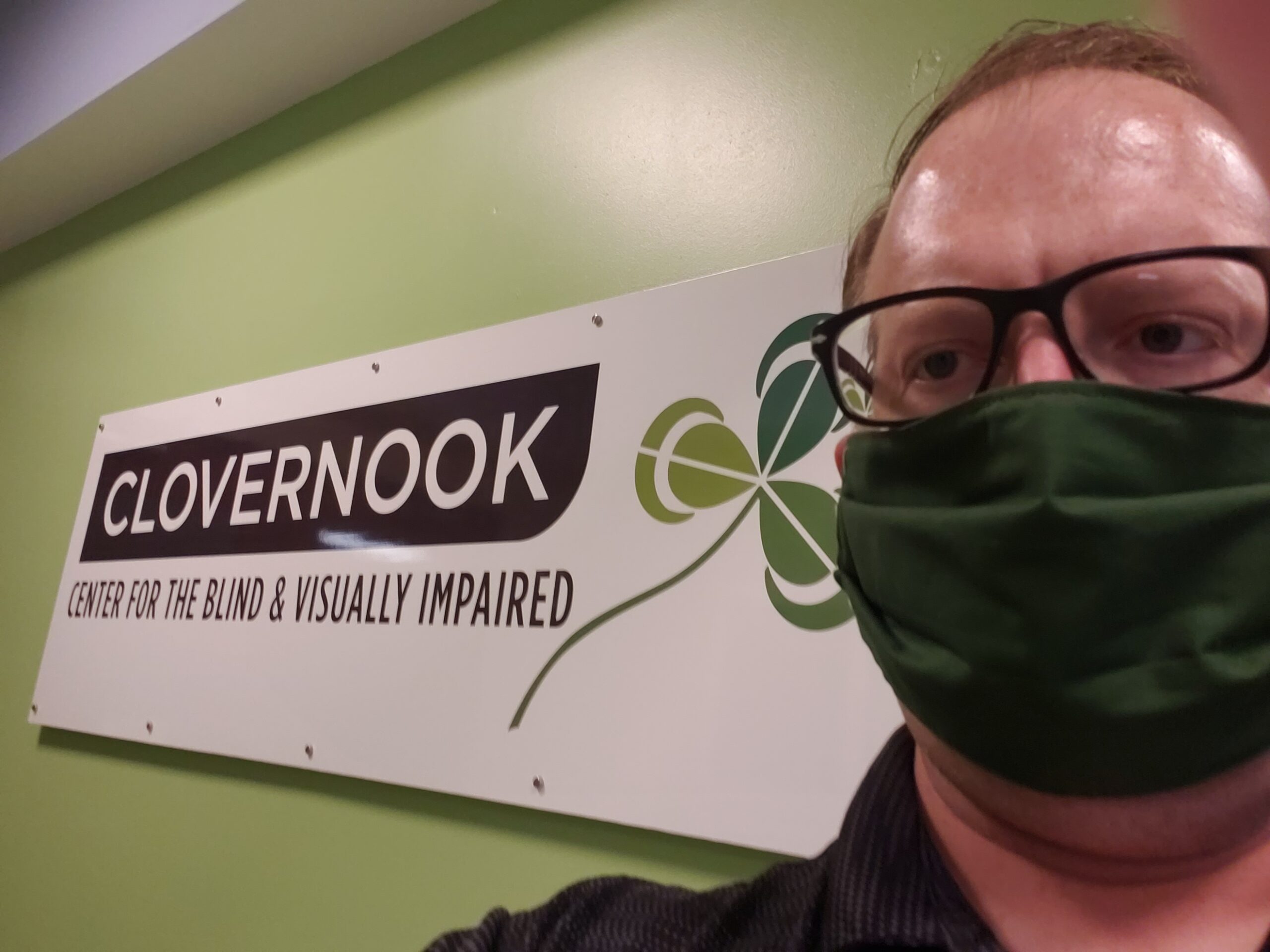Photo of Isaac standing in front of a Clovernook Center sign wearing a green mask and glasses