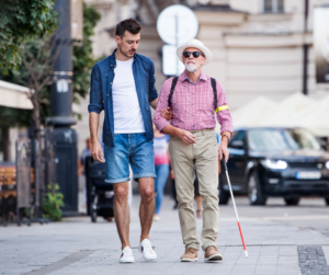 Picture of two men walking down the street one is blind and using a white cane and the other is helping to guide him