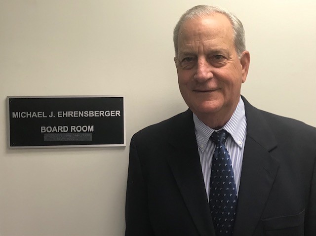 Image of board president, Mike Ehrensberger, standing next to a plaque that reads "Mike J. Ehrensberger Board Room"