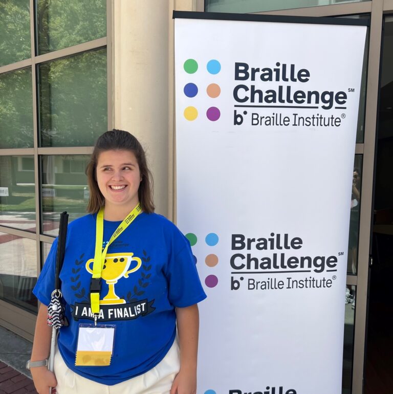 A teen girl wearing a blue t-shirt stands in front of a sign that says Braille Challenge.