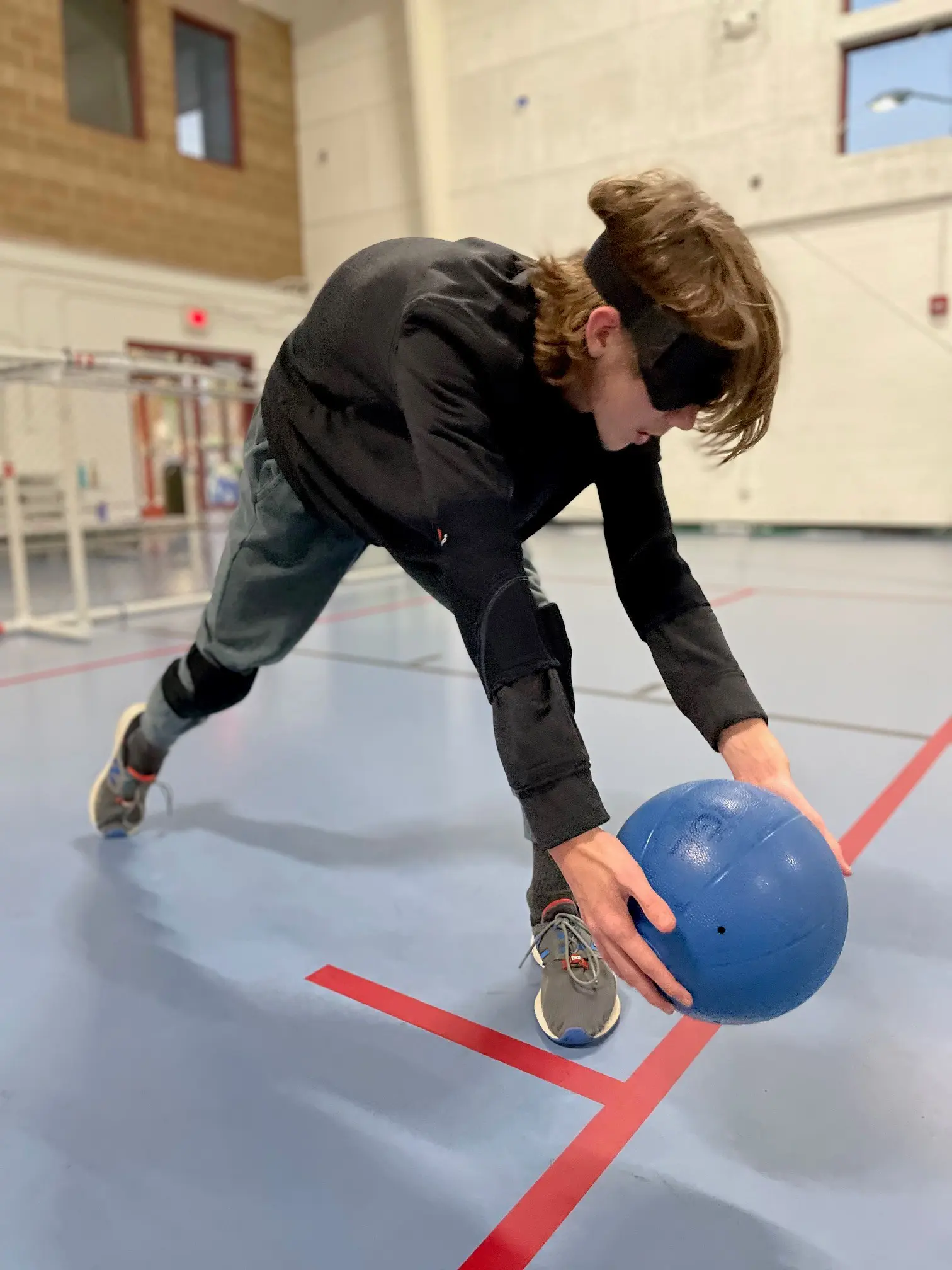 A boy wearing a black long -sleeved shirt and an eye covering lunges for a blue goal ball.