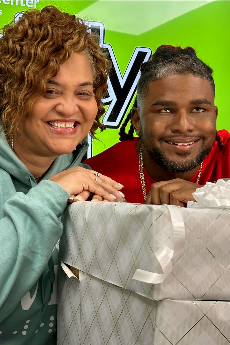 Stephanie Jones and Brian Anderson rest their arms on a large gift wrapped in gray and silver-patterned wrapping paper while on the set of “Boldy BVI.”