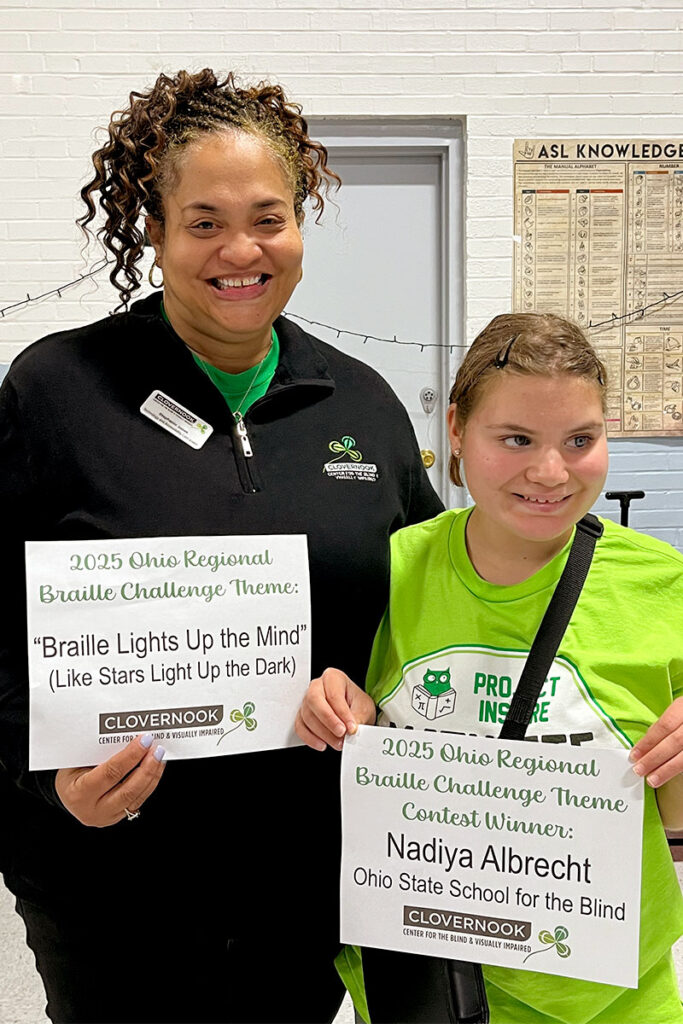 Stephanie Jones stands next to Nadiya Albrecht as they hold up signs announcing the winning 2025 Ohio Braille Challenge Theme.