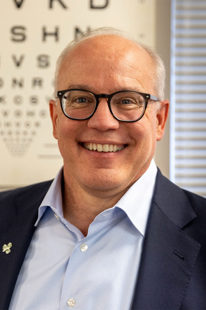 Chris Faust, wearing glasses and dressed in a blue suit, flashes a smile while standing in front of an eye chart.