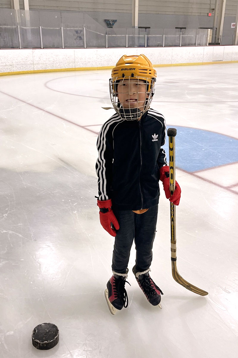 Otto, wearing a hockey helmet and holding a hockey stick, flashes a smile on the ice while standing in front of an adaptive puck.