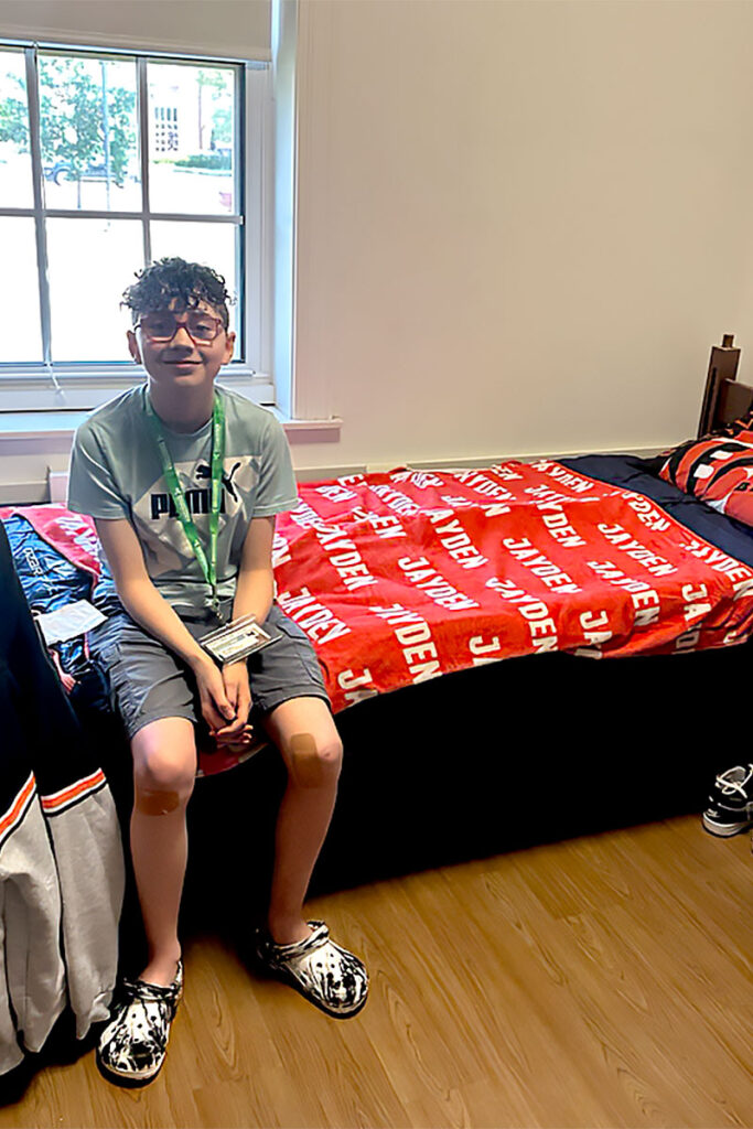 Jayden, wearing glasses and a green Clovernook lanyard with a Camp Abilities nametag, smiles and sits at the end of his bed inside a Miami University dorm room.