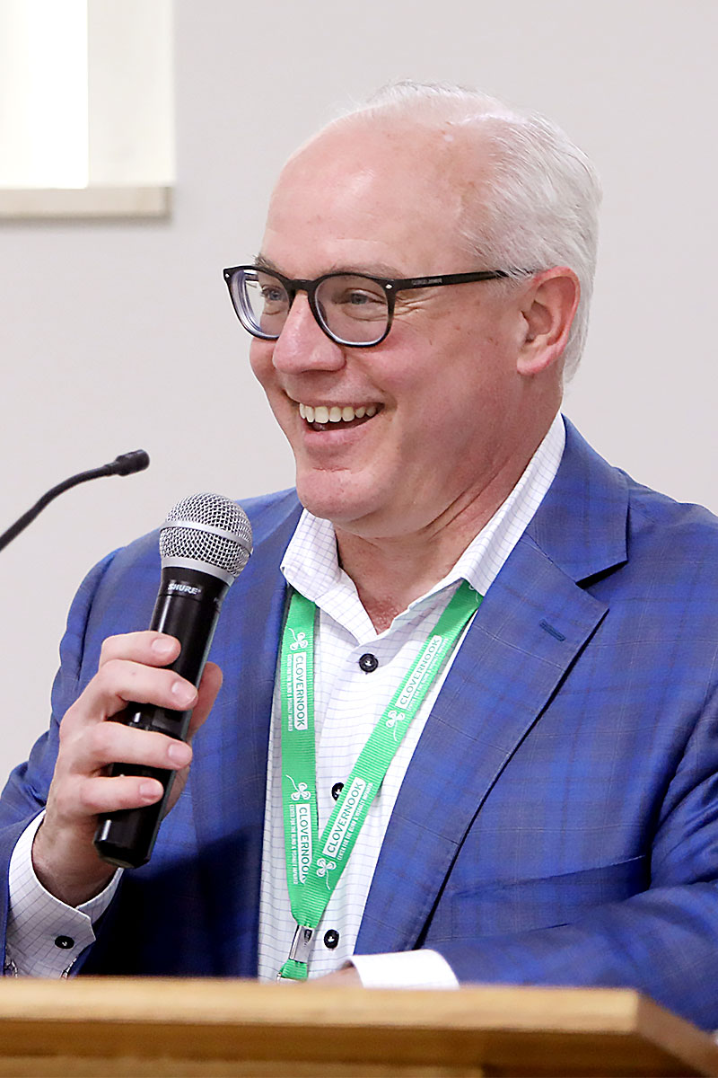Chris Faust, wearing glasses and dressed in a blue plaid suit, speaks into a microphone at a podium.