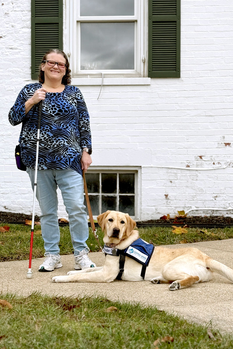 Deanna, holding a white cane in one hand and a leash in the other, smiles as she stands outside Cary Cottage with Buttercup laying in front of her.