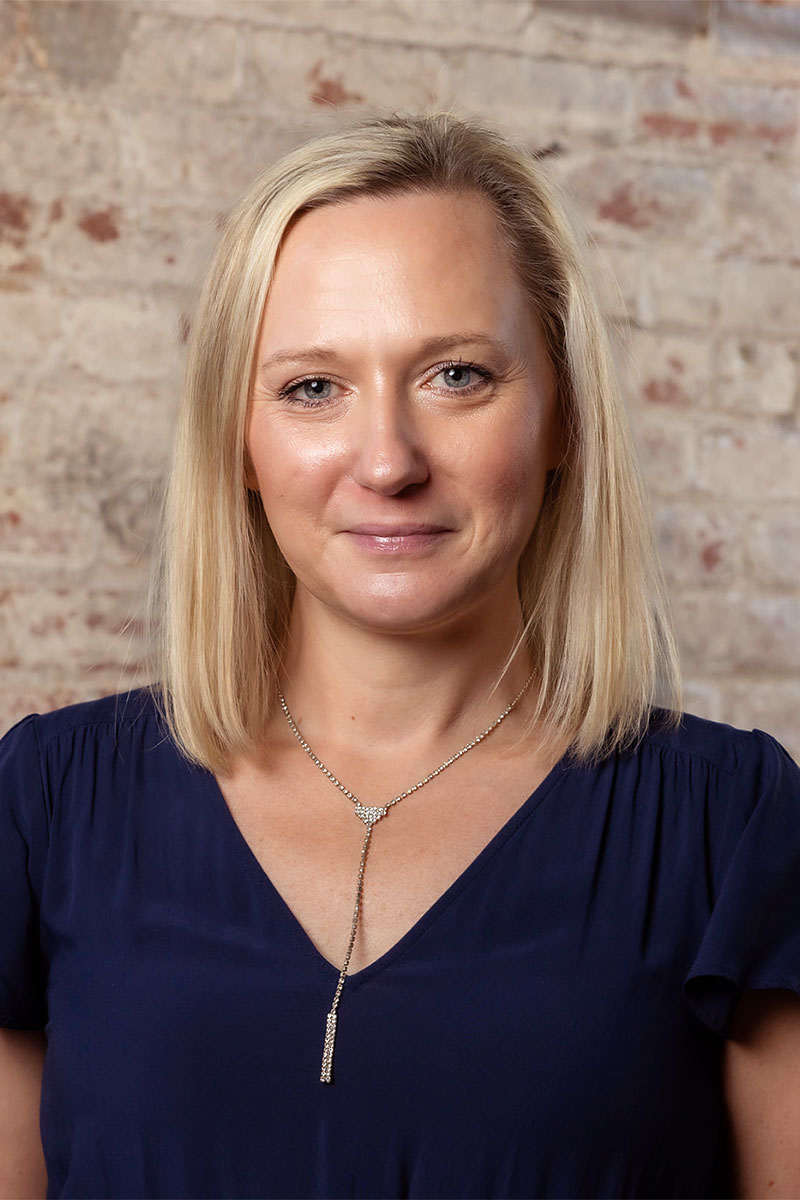 Katie Brass has blond hair and is posing in front of a brick wall, wearing a navy blue shirt and a gold necklace.