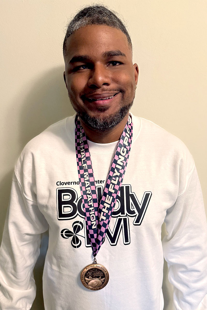 Brian Anderson smiles at the camera while wearing a white long-sleeved Boldly BVI t-shirt and his Flying Pig finisher’s medal.
