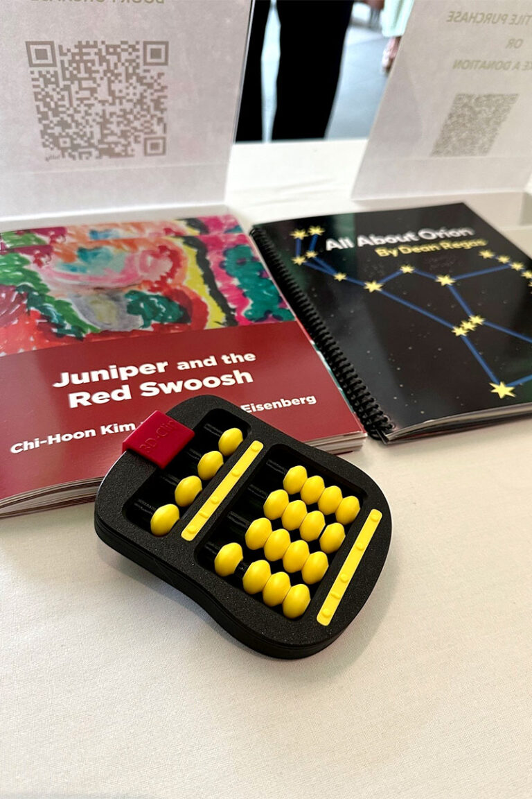 A book with a red cover sits on a table next to a book with a black cover and stars on the front. A black and yellow abacus is placed on top of the book with the red cover.