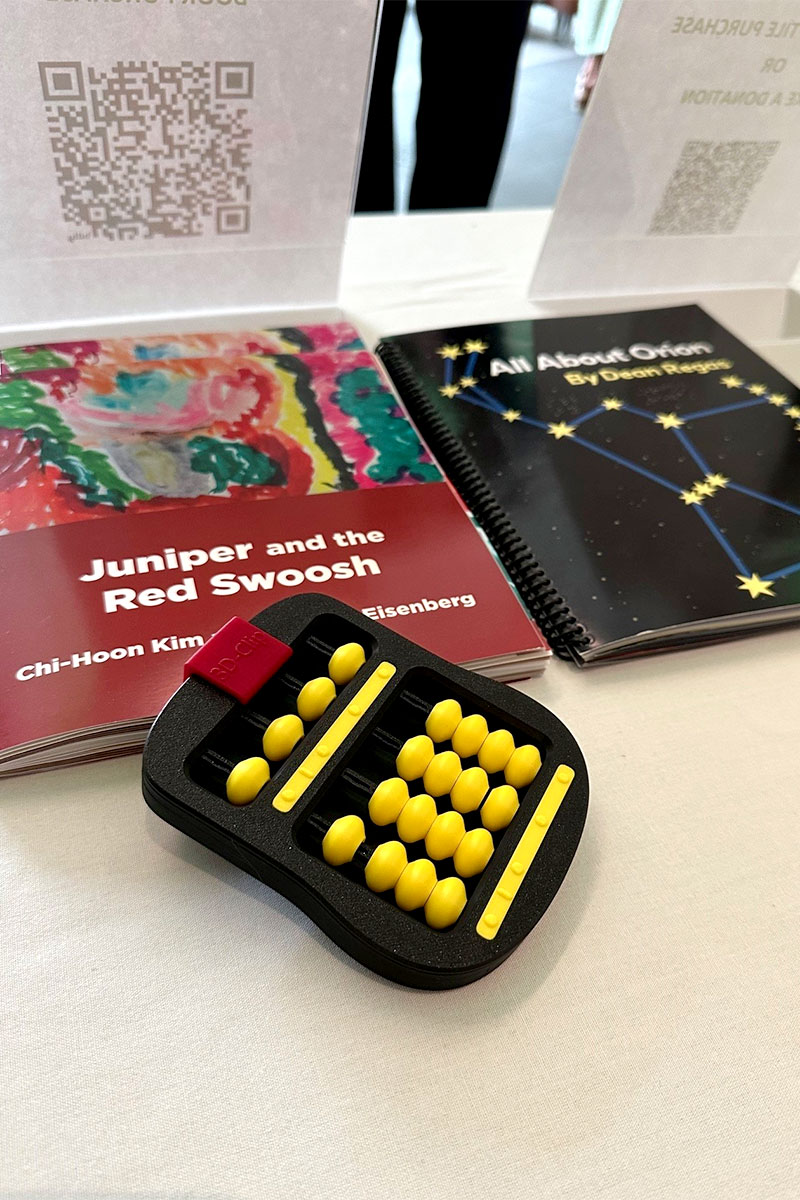 A book with a red cover sits on a table next to a book with a black cover and stars on the front. A black and yellow abacus is placed on top of the book with the red cover.