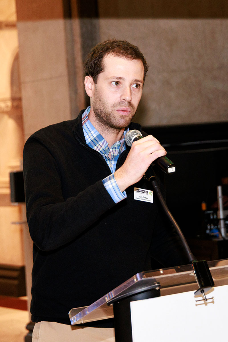 Sam Foulkes has brown hair and is wearing a blue, long-sleeved sweater as he stands at a podium, holding a microphone to address a crowd of people.