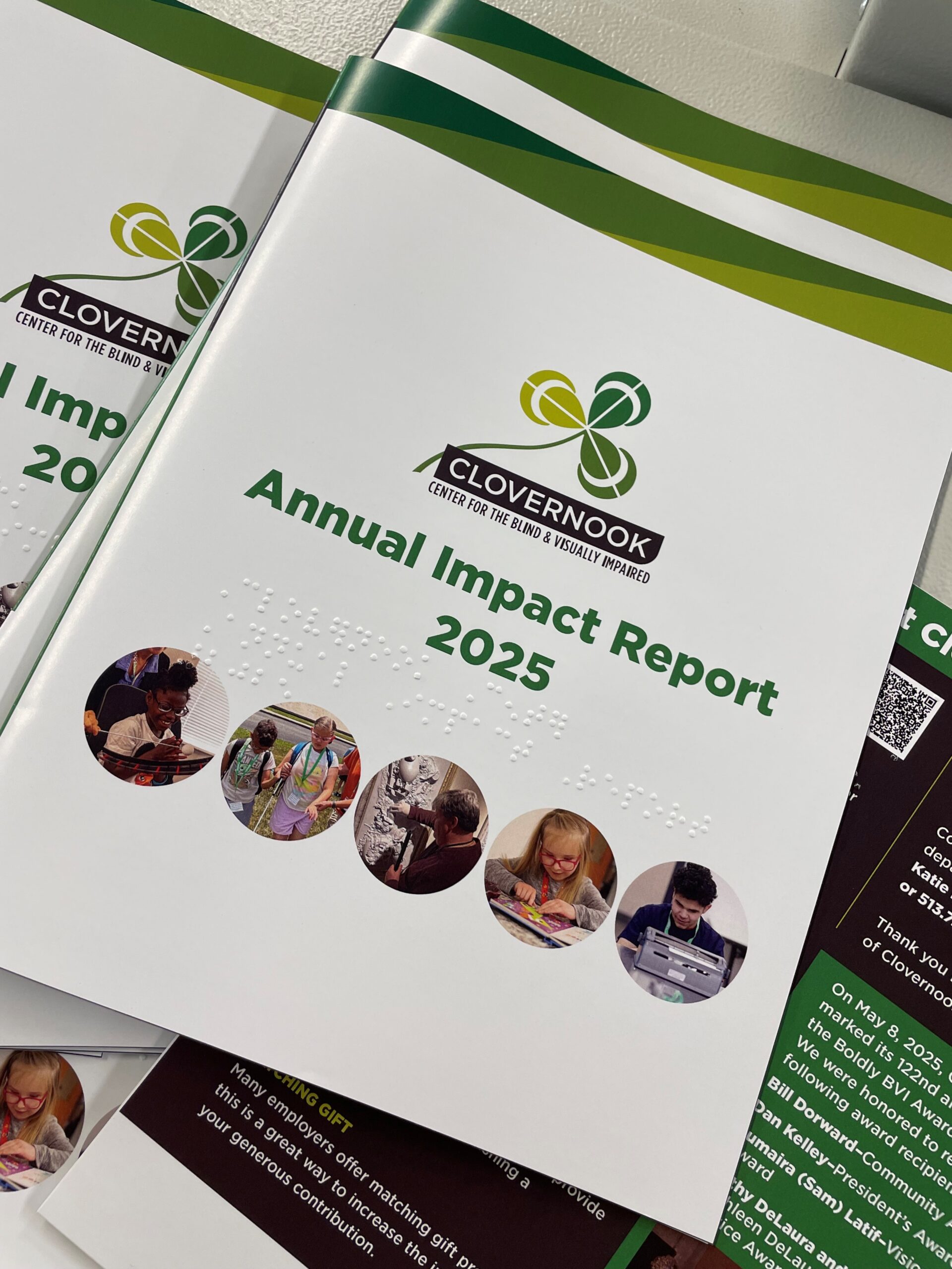 A copy of the 2025 Annual Impact Report sits on top of multiple other copies. The cover reads "Annual Impact Report 2025" and has braille embossed.